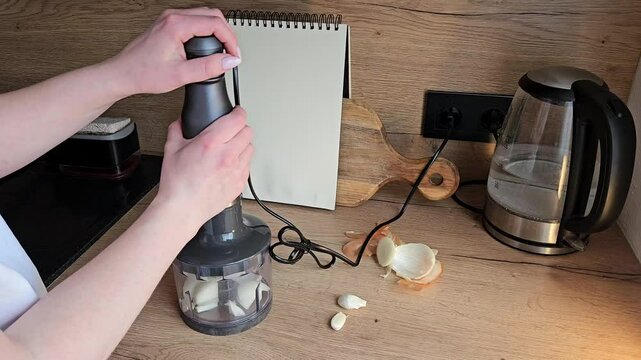 Hands operate a garlic press in a contemporary kitchen setting, processing fresh garlic cloves. A notebook, cutting board
