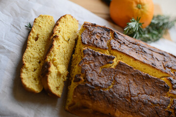 A freshly baked orange cake with two slices cut, placed on a rustic surface. Surrounding the cake are fresh rosemary sprigs and a whole orange, adding a fragrant touch to the scene