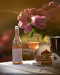 Open bottle of pink wine with white label on holiday table. Pink wine glass. Cake, flowers. Wine tasting. Alcoholic beverage. Winemaking, winery, vineyard. Outdoor setting. Drink concept. Copy space.