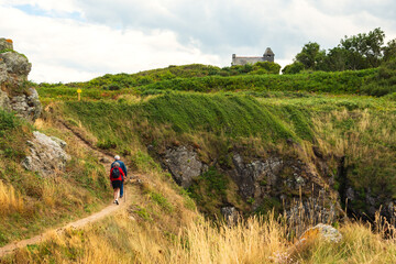Senior couple (back view) hiking in summer at picturesque hills toward a chapel in Emerald Coast, Brittany, France. Healthy active lifestyle, elderly wellbeing concept. French rural tourism background