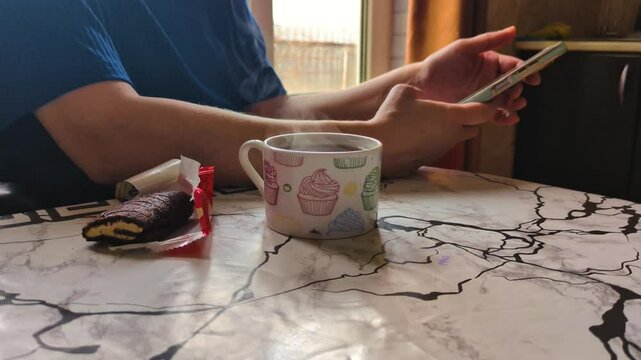 On the marble table there is a mug with a hot drink, from which steam rises. There is an open package of chocolate chip cookies nearby. A man in a blue T-shirt holds a smartphone in his hands while si
