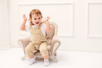 cute toddler sits comfortably on a small vintage-style armchair in a bright, elegant room. The...