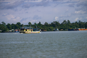 Fototapeta premium A colorful fishing boat sails on a calm river with lush green trees and buildings in the background. Other boats are docked along the shore under a partly cloudy sky