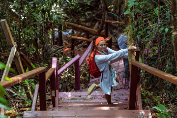 Fototapeta premium A woman with a red headscarf and casual outfit climbs a red wooden staircase in a lush tropical forest, holding the railings. Sunlight filters through the dense green foliage, creating a vibrant atmos