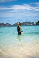 A woman in a wetsuit and headscarf stands in crystal-clear tropical waters, stretching with arms raised. The turquoise ocean, lush green island, and bright blue sky create a serene and scenic paradise