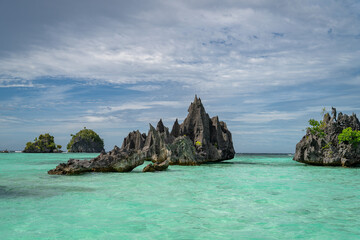 Jagged black limestone formations rise from turquoise waters, creating a dramatic seascape. A natural rock arch adds to the rugged beauty. Greenery clings to the cliffs under a bright blue sky