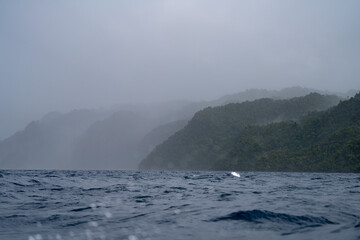 A storm approaches Misool Island in Raja Ampat, with dark clouds over the sea. Waves crash, and raindrops blur the view of the misty jungle-covered coastline, creating a dramatic, moody scene.