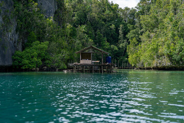 Obraz premium Rustic wooden stilt house stands over emerald waters, surrounded by lush rainforest and towering limestone cliffs, embodying remote island living and tropical serenity.