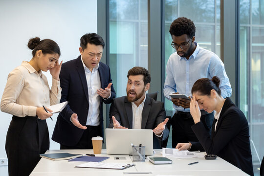 A diverse group of colleagues express frustration around a table with a laptop, showing disappointment with project outcomes and facing challenges