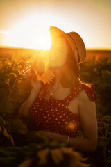 A beautiful young woman in a red polka-dot dress and straw hat smiles, holding a sunflower in a golden field at sunset. Her glowing skin and soft curves contrast with the warm light and green leaves