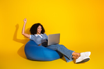 Young woman in casual attire happily using a laptop while sitting on a beanbag chair on a vibrant...