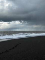 Footprints on black sand beach with ocean waves under storm clouds