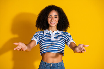 Cheerful young woman with outstretched arms in front of vivid yellow background, wearing a trendy...