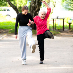 Obraz premium Smiling grandmother and granddaughter doing sports together in the park. Old and young women do a warm-up. They walk along the sidewalk, bouncing.