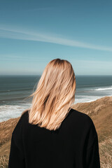 Fototapeta premium Blonde woman standing on coastal cliffside overlooking the sea, peaceful minimal lifestyle scene with clear blue sky
