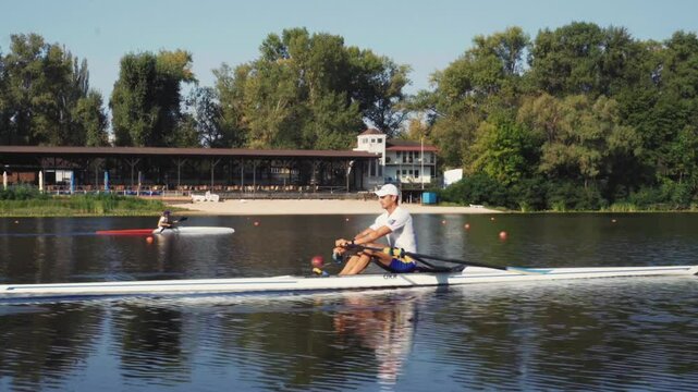 Rower solo training. Side view of young caucasian male rower, during a rowing practice, athlete sitting in a boat in the river Dnipro, rows through a calm water in autumn. 4k footage. Caucasian man tr