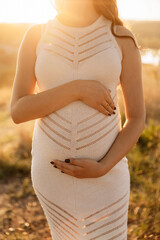 Close-up photo of a pregnant girl in a dress against a summer sunset. Future parenthood