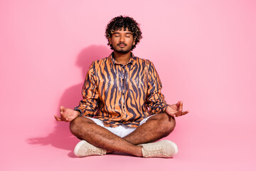 Young stylish man with curly hair meditating cross-legged against a vibrant pink background wearing...