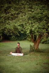 Man, yoga and lotus for meditation by tree, eyes closed and spiritual on grass in morning at park. Mature person, zen and peace in garden with balance, breathing and mindfulness on ground in France