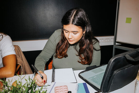 Young woman writing on diary while sitting with laptop in classroom at university
