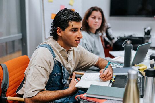 Young man wearing bib overalls doing homework while sitting near table in classroom at university