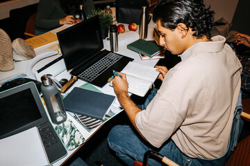 High angle view of left handed young man writing on diary while studying with laptop in classroom at university