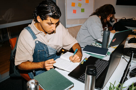 Left handed young man writing on diary while studying with laptop in classroom at university