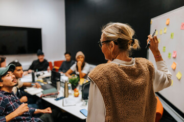Rear view of female teacher holding felt tip pen teaching multiracial students in classroom at college