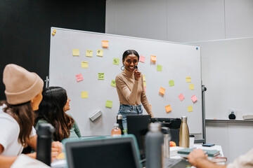 Happy young female student standing near whiteboard while discussing with friends during project meeting in classroom at
