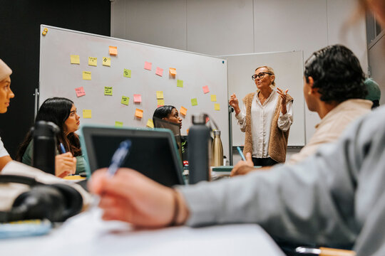 Female professor explaining while teaching students and standing near whiteboard in classroom at university