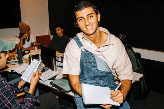 Portrait of happy young man wearing bib overall holding diary while sitting on table in classroom
