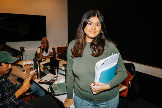 Portrait of smiling young female student holding books while standing near table in classroom at college