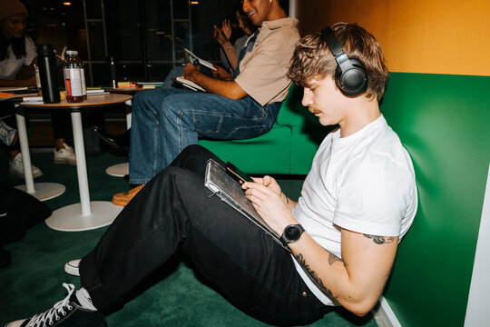 Full length of young man with tattooed arm using smart phone and wearing wireless headphones while leaning on wall in un
