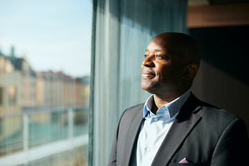 Thoughtful smiling bald male entrepreneur looking through window at office