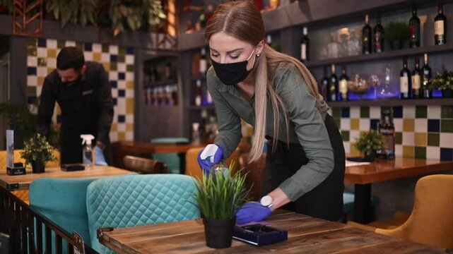 Young restaurant waiters cleaning and disinfecting tables and surfaces during Coronavirus pandemic disease. They are wearing protective face masks and gloves.