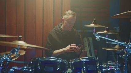 Drummer takes a break during practice to check messages on his phone while surrounded by drum kit and studio lights. Focus on dedication to music and modern interruptions.