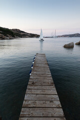 Wooden pier and sailboats sailing in evening calm sea of marvellous Porto Rafael, Costa Smeralda, Sardinia, Italy. Symbol for relaxation, wealth, leisure activity.
