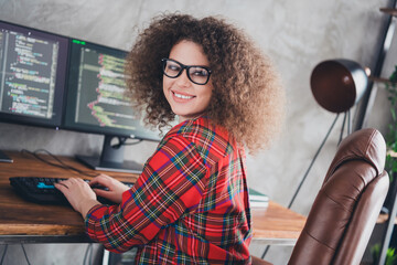 Young female programmer working from home in a casual checkered shirt, coding on dual monitors in a modern office setup