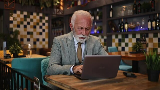 Handsome senior businessman sitting at restaurant table and working something on his laptop computer. It is obvious that he have some business problems and he is very worried.