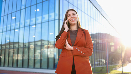 Fototapeta premium Businesswoman smiling while talking on phone in front of modern office building