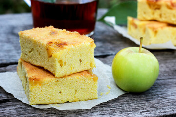 A slice of charlotte pie rests on a wooden table against a green apple and a glass of tea. Home baking on a summer afternoon in the garden