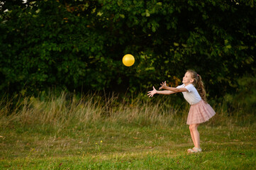 Little girl playing happily with ball on grass in park on sunny day, throwing and catching it. Children's games on vacation, playing ball outside