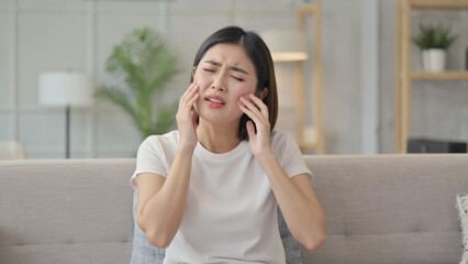 Agonizing Pain: An Asian woman, captured indoors on a couch, expresses discomfort while touching her face, suggesting a sudden onset of pain or ache. 