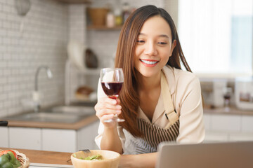 Young woman enjoying glass of wine while chatting through video call in cozy home kitchen
