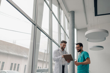 Two doctors smiling and using a tablet in hospital corridor