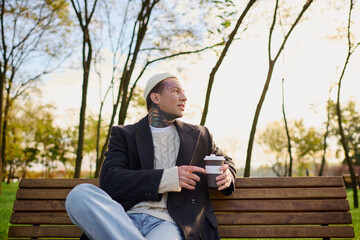 Fototapeta premium Stylish young man with tattoos enjoys a coffee in a serene park setting during golden hour