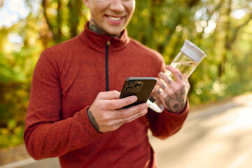 Stylish young man enjoying a refreshing drink while using his smartphone in a sunlit park