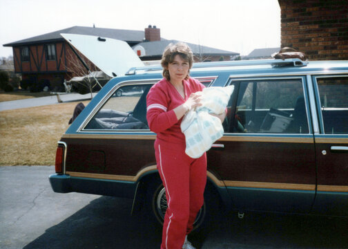 Circa 1985 - Vintage family photo of new mom bringing her baby home from hospital, posed outside of a wood paneled station wagon. Vintage scan, may have imperfections