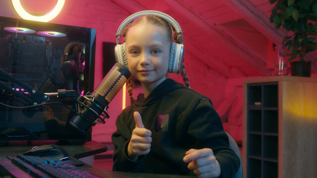 Caucasian little teen girl daughter child kid streamer gamer influencer gen z video internet game computer addiction at night radio hold microphone put on headphones prepare to stream showing thumb up