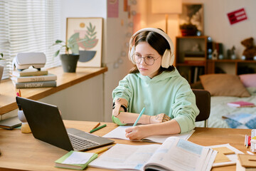 Young girl with prosthetic hand wearing headphones working at cluttered desk with books, laptop